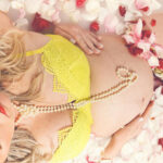 Quelle lingerie de grossesse choisir pour une petite poitrine ? 6 Femme enceinte portant un ensemble de lingerie jaune et des perles, allongée dans un bain de lait parsemé de pétales de roses roses et blanches.