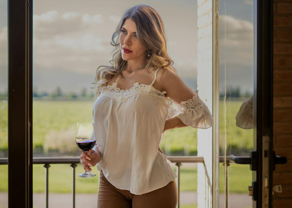 On arrête le mensonge : ces hauts ne sont PAS réservés aux fortes poitrines 7 Une femme élégante tenant un verre de vin rouge sur un balcon, profitant d'un moment de calme et de sérénité face à un paysage naturel.