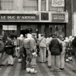 Une foule réunie devant un établissement nommé "Le Duc d’Artois", illustrant un moment de vie sociale et de communauté.
