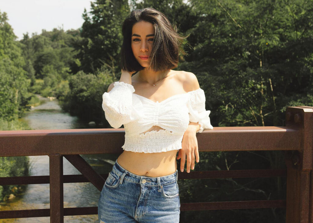 Justaucorps, crop top, débardeur : comment le porter haut la tête même sans volume 7 Une femme sereine portant un haut blanc et un jean, s'appuyant sur une rambarde dans un cadre naturel, illustrant l'élégance et l'acceptation de soi.