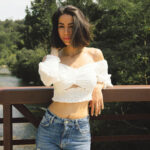 Justaucorps, crop top, débardeur : comment le porter haut la tête même sans volume 9 Une femme sereine portant un haut blanc et un jean, s'appuyant sur une rambarde dans un cadre naturel, illustrant l'élégance et l'acceptation de soi.