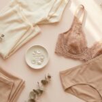 Coton, Microfibre, Dentelle : Quelle matière de culotte choisir pour allier confort et hygiene intime ? 11 Composition flat-lay esthétique et épurée présentant une culotte en dentelle beige nude, des vêtements en maille pliés de couleur neutre, une coupelle de boutons rétro et des branches d'eucalyptus séchées sur une surface pastel avec des ombres douces.